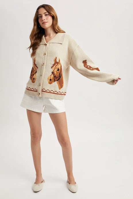 A woman poses against a plain light background wearing the Zertz HORSE GRAPHIC BUTTON CARDIGAN, featuring bold horse head designs, styled with white shorts and white shoes.