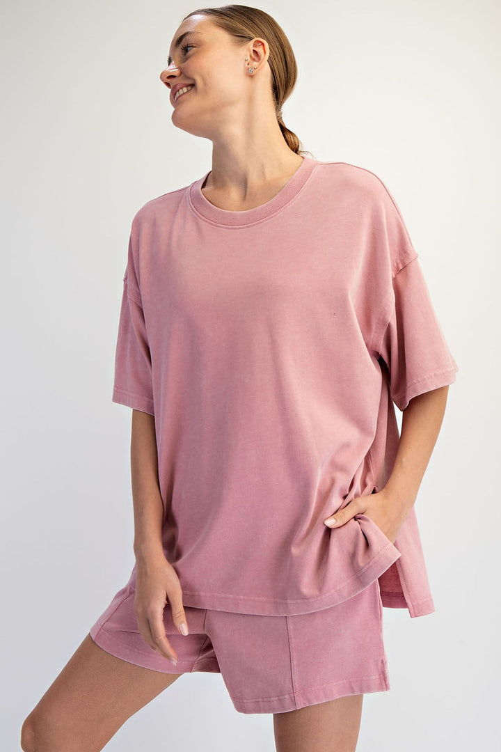 Someone wearing The Mellow Yellow Set by Loca on Main—a loose mineral washed tee and relaxed shorts—stands against a plain light background, smiling to the side with one hand in their pocket—perfect everyday loungewear.
