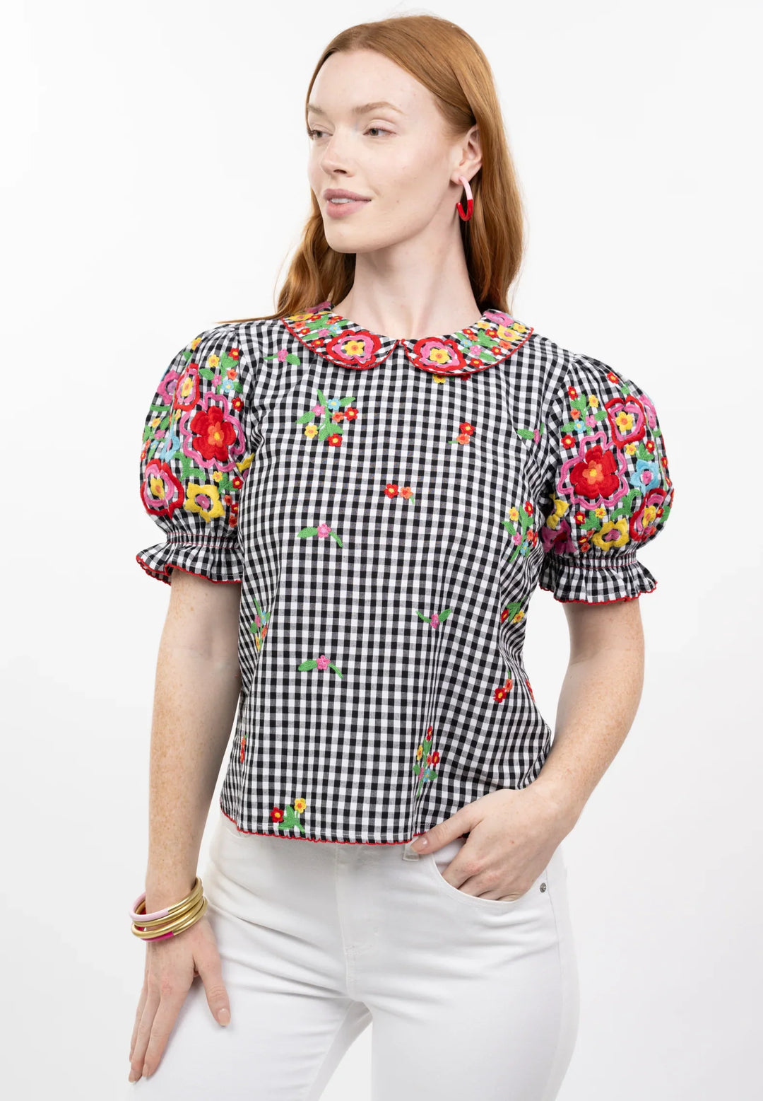 A woman with long red hair models the Zertz GINGHAM GARDEN TOP, a black and white checkered blouse with a bib collar, colorful embroidery, puffed sleeves, and scalloped edges. She pairs it with white pants and gold bangles against a white background.