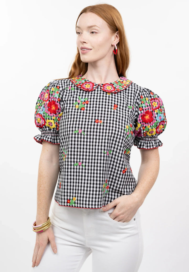 A woman with long red hair models the Zertz GINGHAM GARDEN TOP, a black and white checkered blouse with a bib collar, colorful embroidery, puffed sleeves, and scalloped edges. She pairs it with white pants and gold bangles against a white background.