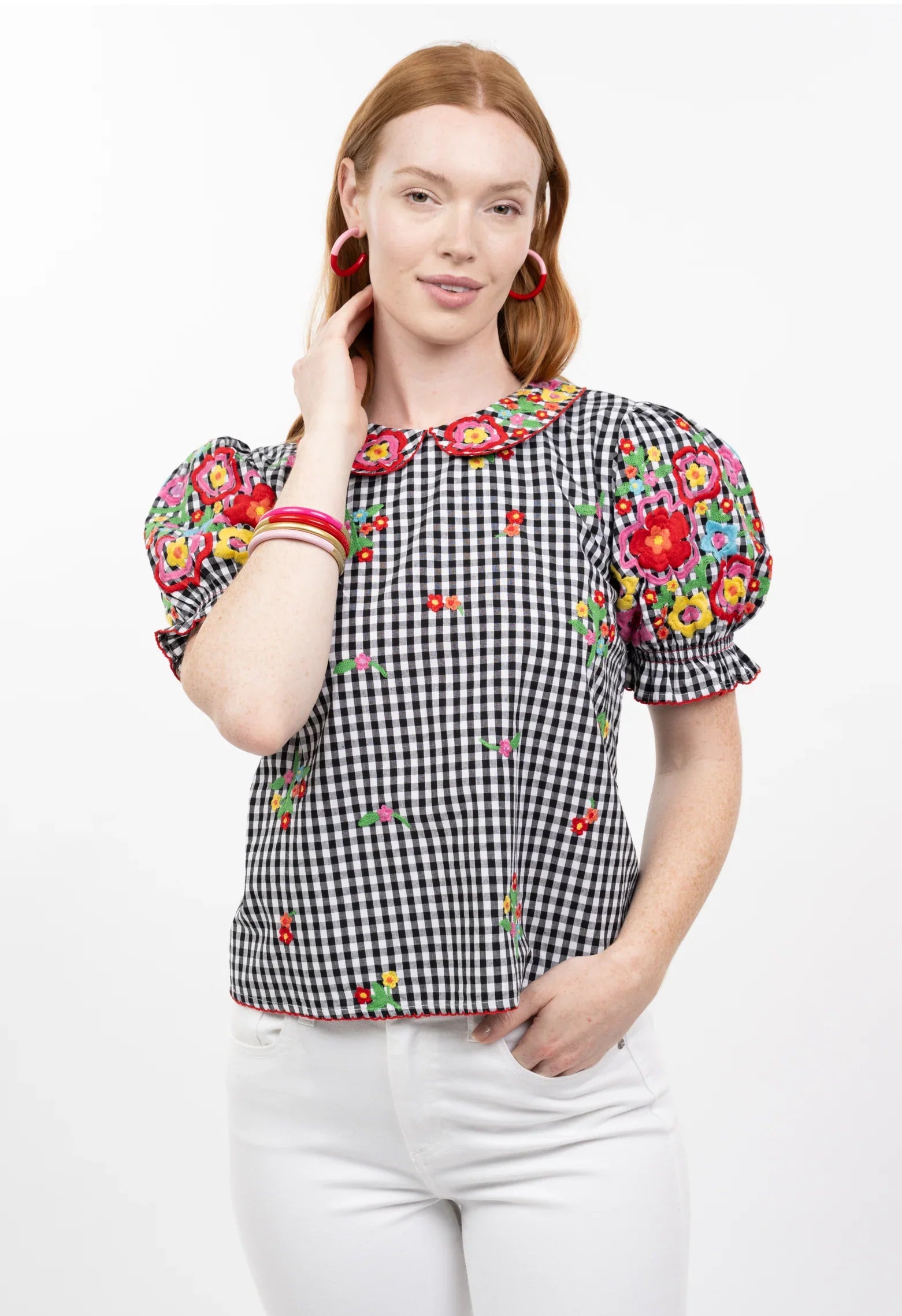 A woman with long red hair models the Zertz GINGHAM GARDEN TOP, featuring black and white gingham, heavy embroidery, puffed sleeves, and a scalloped collar. She pairs it with white pants and pink hoop earrings against a plain white background.