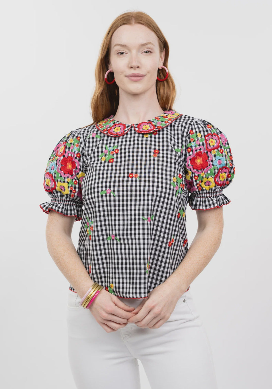 A woman with long red hair wears the Zertz GINGHAM GARDEN TOP, a black and white checkered blouse with colorful floral embroidery on the sleeves and bib collar, paired with white pants. She smiles softly before a plain white background.