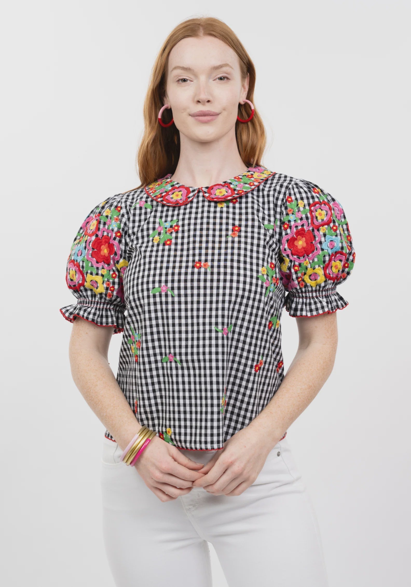 A woman with long red hair wears the Zertz GINGHAM GARDEN TOP, a black and white checkered blouse with colorful floral embroidery on the sleeves and bib collar, paired with white pants. She smiles softly before a plain white background.