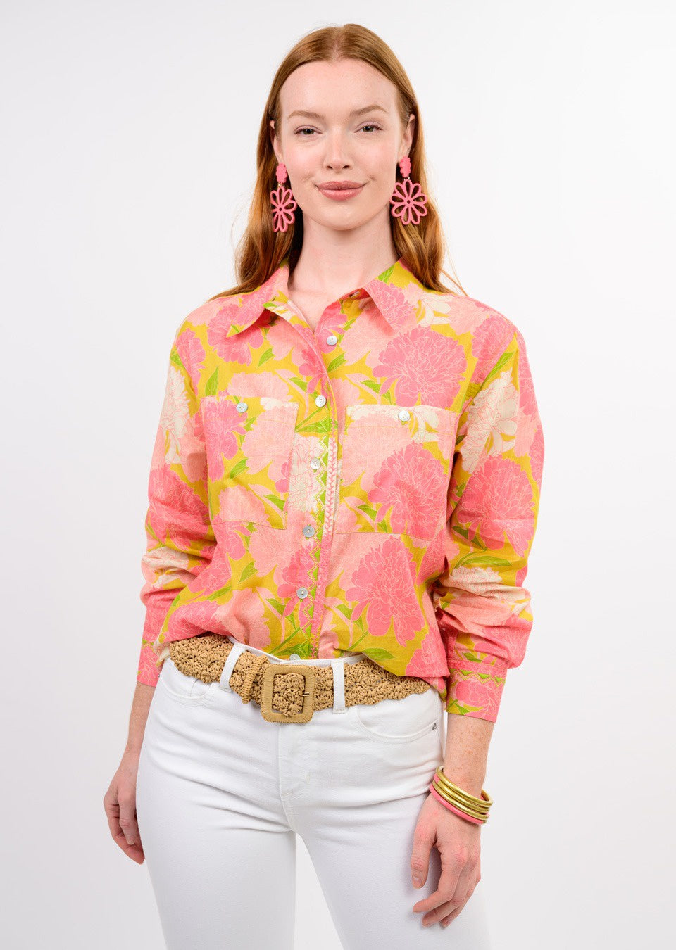 Wearing the Zertz BLOOMS A PLENTY SHIRT, white pants, a woven beige belt, pink flower earrings, and gold bangles, a woman stands against a plain white background with a soft smile.