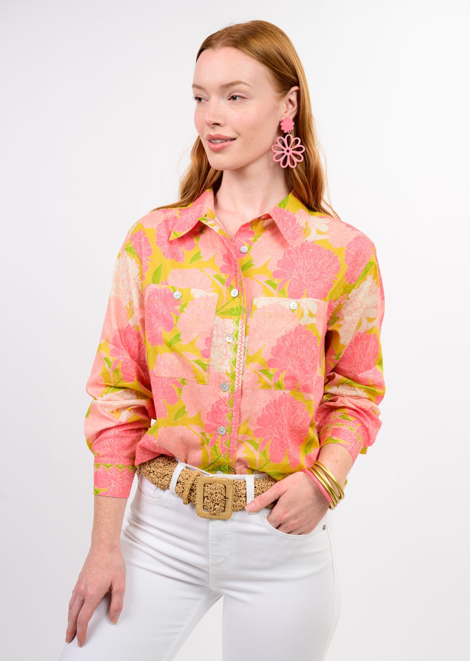 A woman with long red hair wears the Zertz BLOOMS A PLENTY SHIRT—a pink and yellow floral print top—paired with white pants, a woven belt, pink flower earrings, and gold bangles against a plain white background.