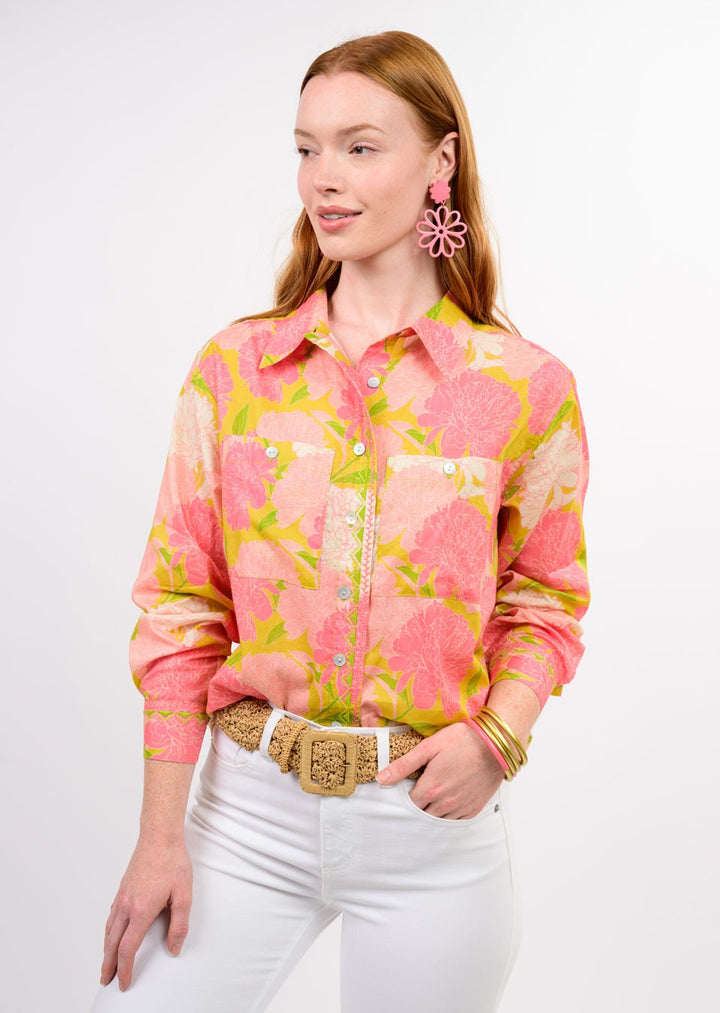 A woman with long red hair wears the Zertz BLOOMS A PLENTY SHIRT—a pink and yellow floral print top—paired with white pants, a woven belt, pink flower earrings, and gold bangles against a plain white background.
