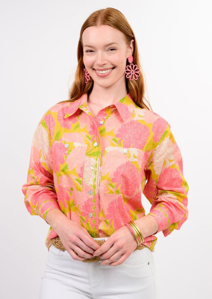 A woman with long red hair smiles, wearing the Zertz BLOOMS A PLENTY SHIRT, a vibrant pink, yellow, and green floral print top. She pairs it with large pink flower-shaped earrings and gold bracelets, set against a crisp white background.