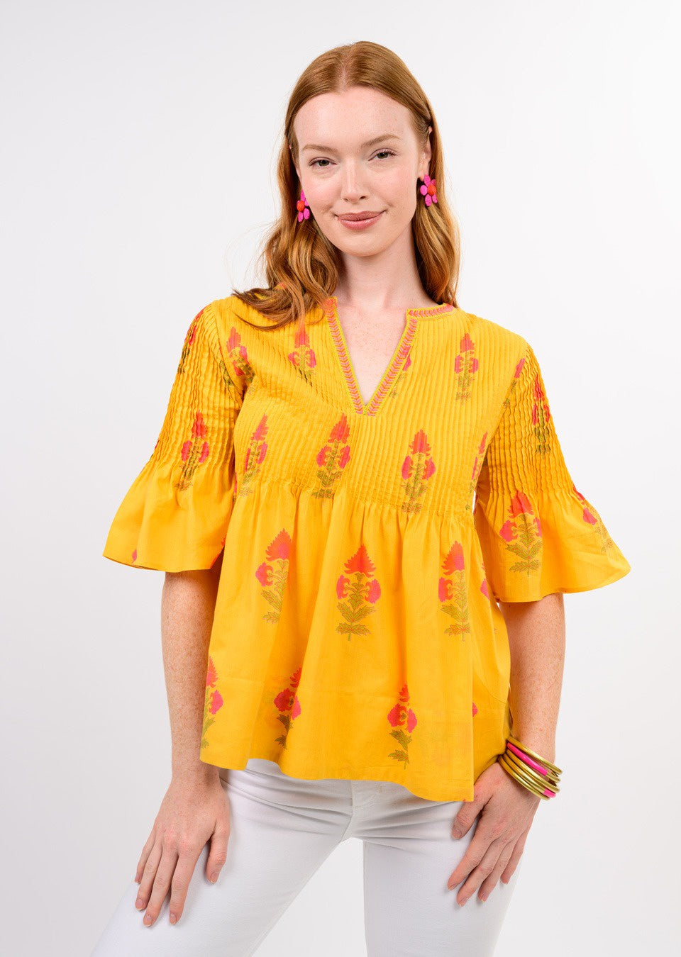 A woman with long red hair models the Zertz BURST OF SUNSHINE TOP—a floral piece with flared sleeves—paired with white pants, pink earrings, and bangles. Posed against a plain white backdrop, it&