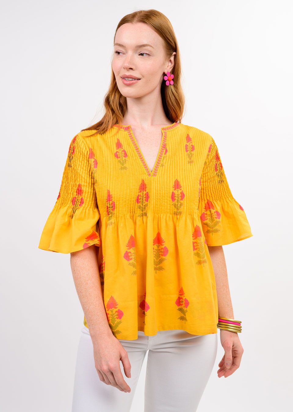A woman with long red hair models the Zertz BURST OF SUNSHINE TOP—a yellow embroidered blouse with red and pink blooms—paired with white pants, pink flower earrings, and colorful bangles against a plain white background.
