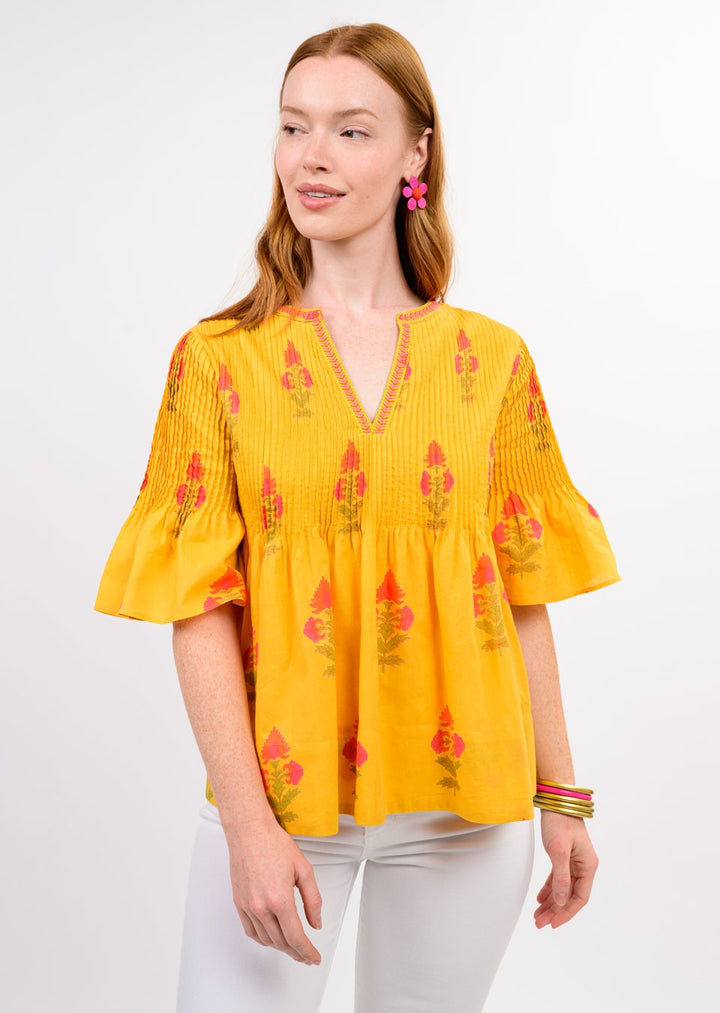 A woman with long red hair models the Zertz BURST OF SUNSHINE TOP—a yellow embroidered blouse with red and pink blooms—paired with white pants, pink flower earrings, and colorful bangles against a plain white background.