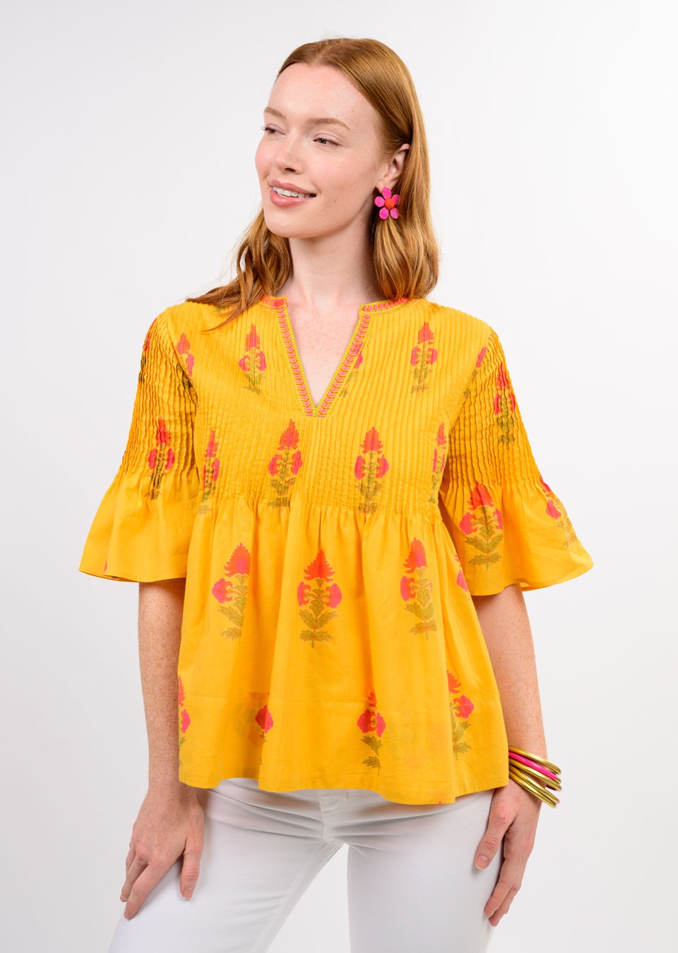 A woman with long red hair smiles in the Zertz BURST OF SUNSHINE TOP, featuring vibrant floral embroidery, styled with white pants and pink earrings—a fresh spring and summer look against a light background.