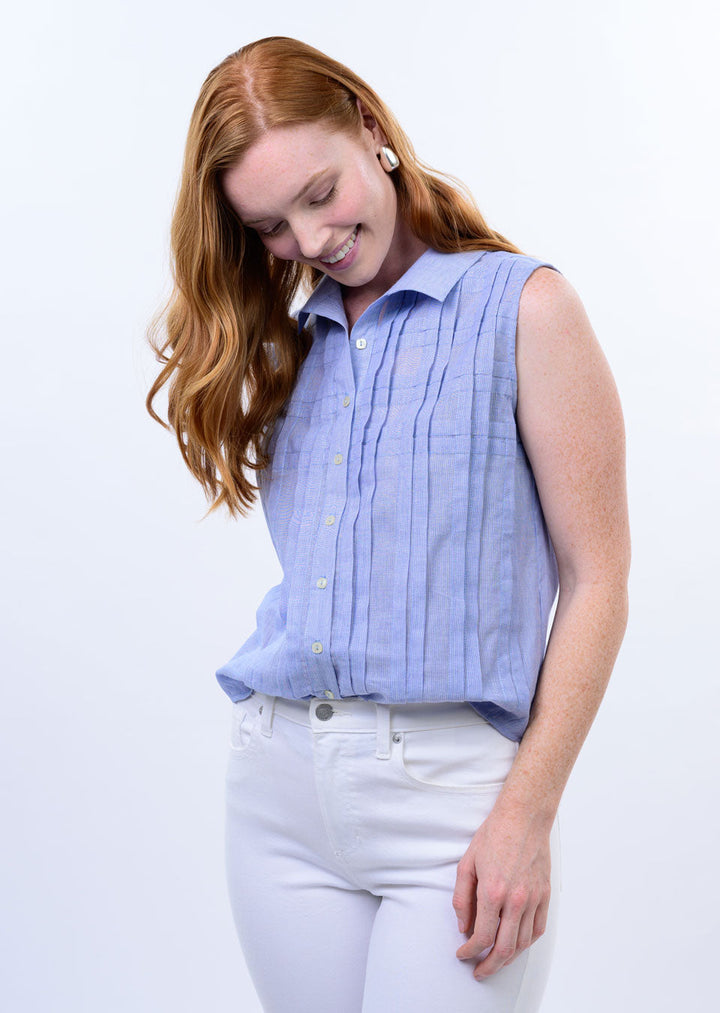 A woman with long red hair smiles while looking down, wearing the Zertz TUCKED PATTERN SHIRT in light blue with vertical pleats and white pants, set against a white background.