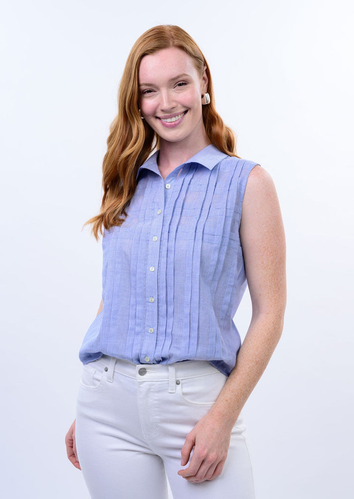 A woman with long red hair smiles at the camera, wearing the Zertz TUCKED PATTERN SHIRT—a sleeveless, tailored-fit, light blue button-up—paired with white pants. She stands against a plain background with one hand in her pocket.