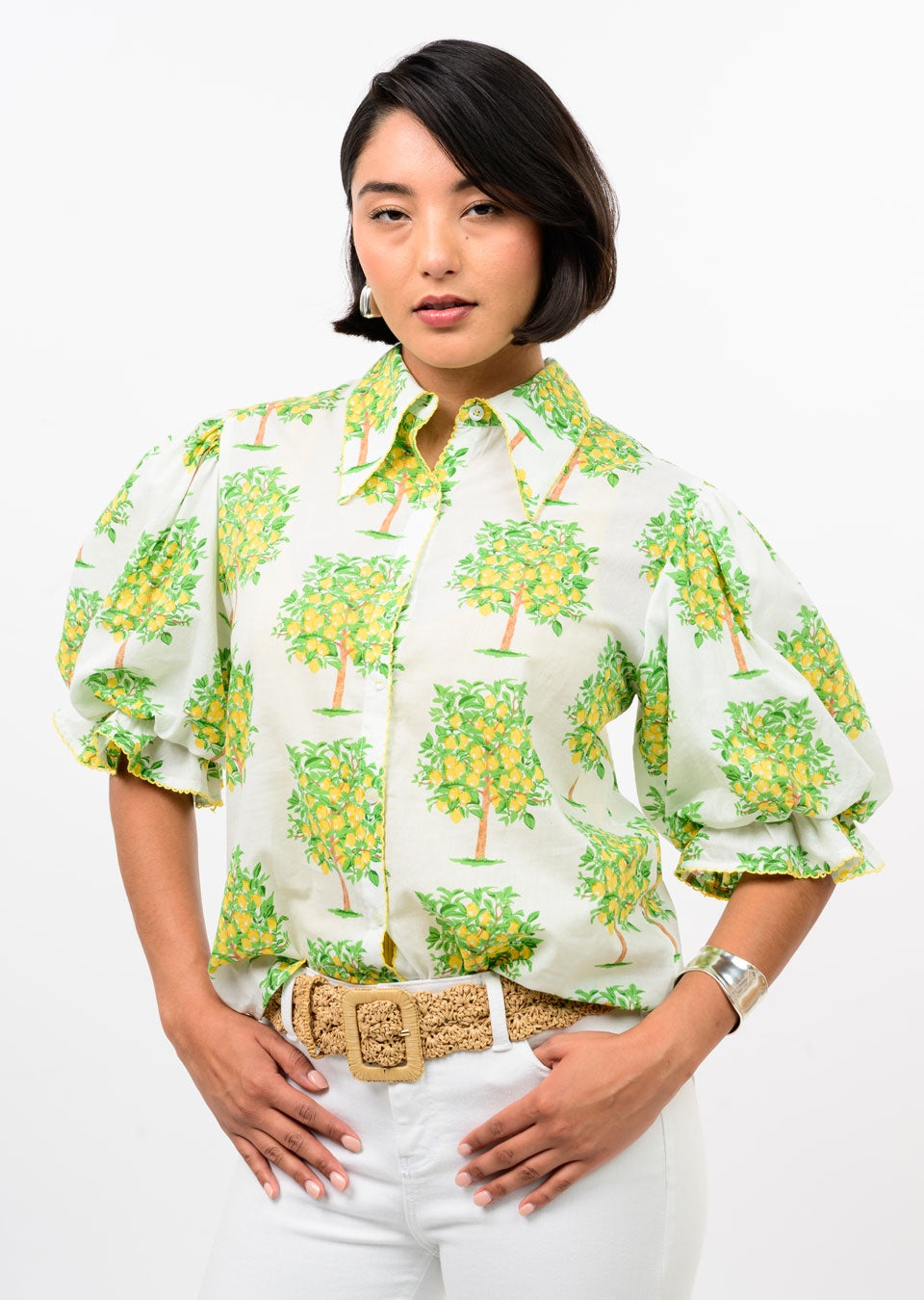 A woman with short dark hair wears the Zertz LEMON TREE TOP—a white blouse with green and yellow tree prints and puffed sleeves—paired with white pants, a woven belt, hoop earrings, and a silver bracelet against a plain white background.