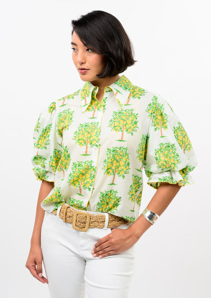 A woman models the Zertz LEMON TREE TOP, inspired by lemons for a fresh spring/summer vibe, paired with white pants, a beige woven belt, and a silver cuff bracelet against a white background.