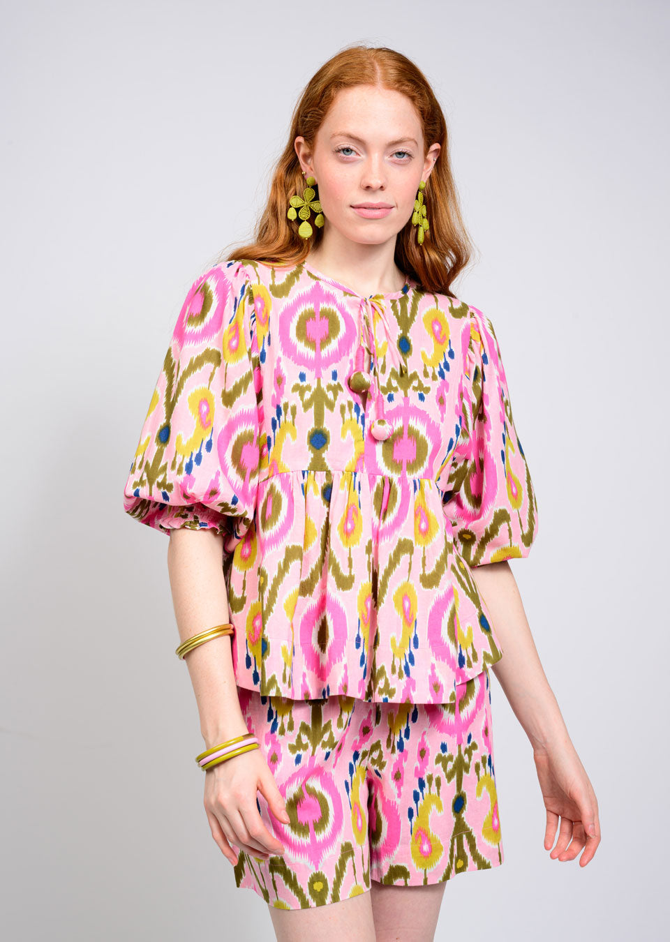 A woman with long red hair wears a pink, green, and white hand-dyed blouse with puffed sleeves and Zertz IKAT SHORT featuring geometric patterns. She accessorizes with green earrings, gold bangles, and stands before a plain light background.