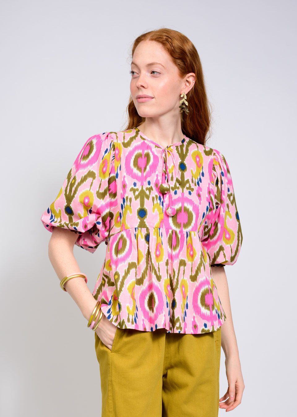A woman with long red hair wears the Zertz IKAT TOP with geometric patterns and puff sleeves, paired with mustard yellow pants. She accessorizes with gold earrings and bracelets, standing against a plain light background.