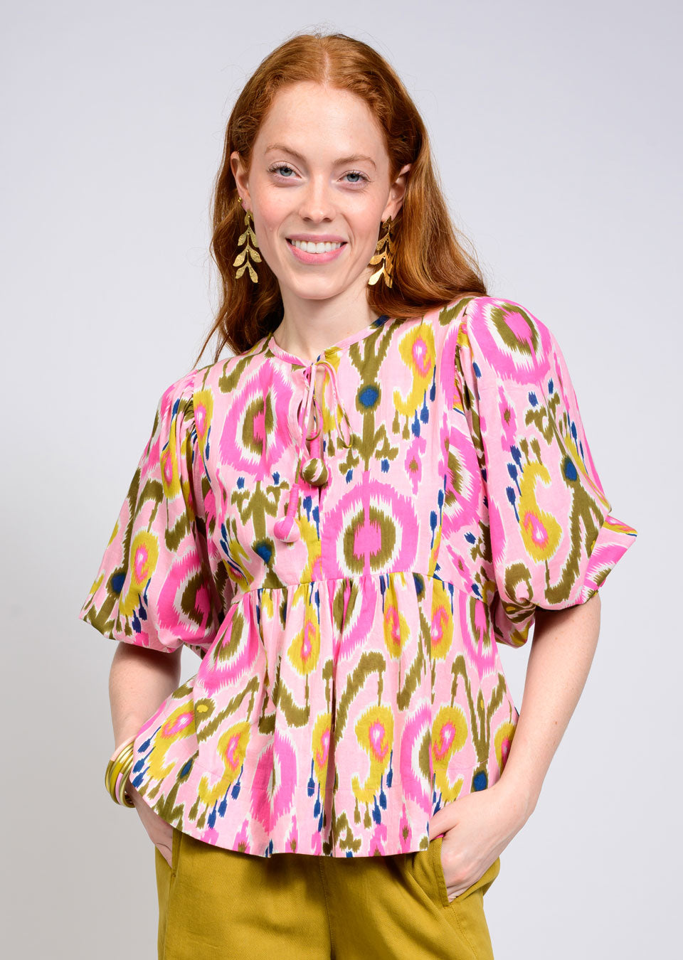 A woman with long red hair smiles, wearing the Zertz IKAT TOP with pink, yellow, and green geometric patterns and short puffed sleeves, paired with olive green pants, gold earrings, and a chunky bracelet for a handcrafted touch.