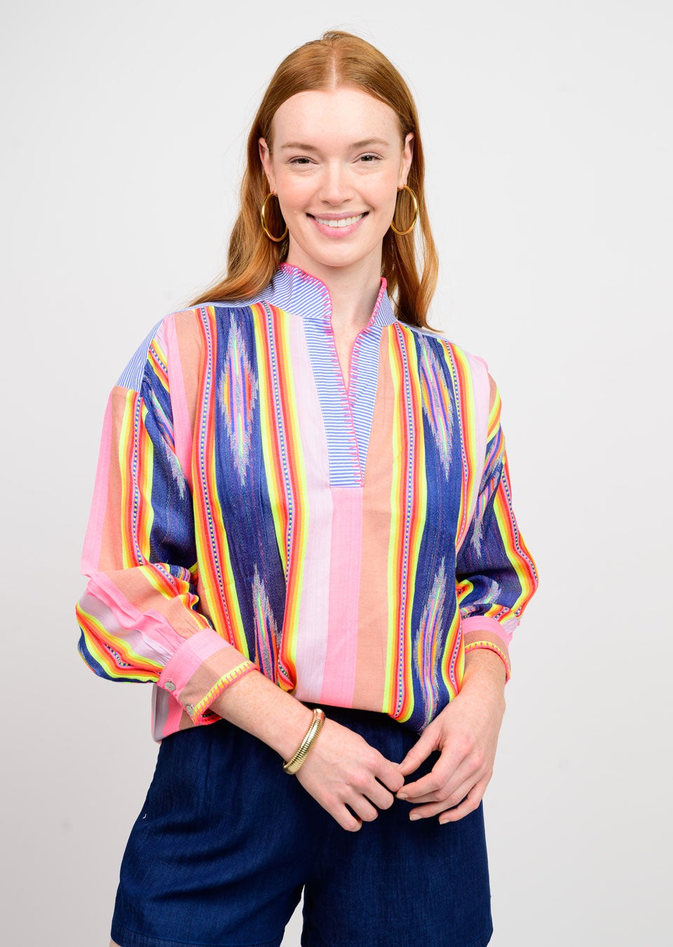A woman with long red hair smiles, wearing the Zertz IKAT STRIPE PULLOVER TOP and navy shorts. She accessorizes with large hoop earrings and gold bracelets, standing against a plain light background.