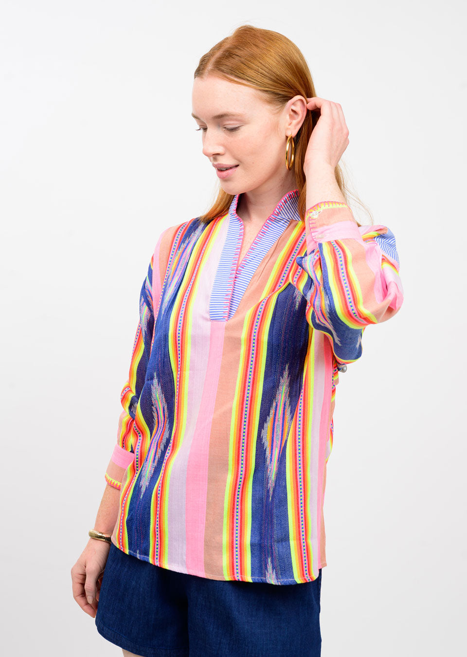 A woman with long red hair wears the Zertz IKAT STRIPE PULLOVER TOP with blue, pink, and orange patterns and dark blue shorts. She looks down, touching her hair, against a plain white background.