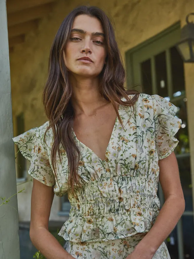 A woman with long brown hair stands outdoors, wearing the SEREIA FLORAL TOP by Zertz—its ruffled sleeves and deep V-neck complementing the rustic building with green-trimmed windows in the background.