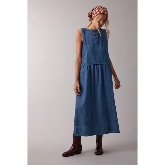 A woman stands against a plain, light background wearing the Zertz SLVLESS MAXI DRESS in blue denim with brown boots and a red patterned headscarf, looking down with one hand by her side.