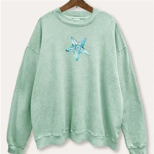 The Blue Starfish Sweatshirt by Loca on Main is a light green oversized top featuring a blue starfish graphic at the center chest, shown on a wooden hanger against a plain background.