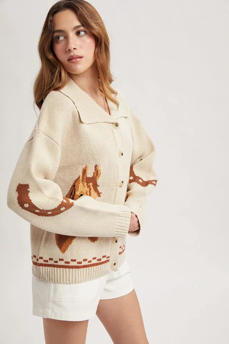 A woman wears the Zertz HORSE GRAPHIC BUTTON CARDIGEN—cream-colored with brown horse patterns on the sleeves and back—paired with white shorts, standing against a plain light background and looking off to the side.
