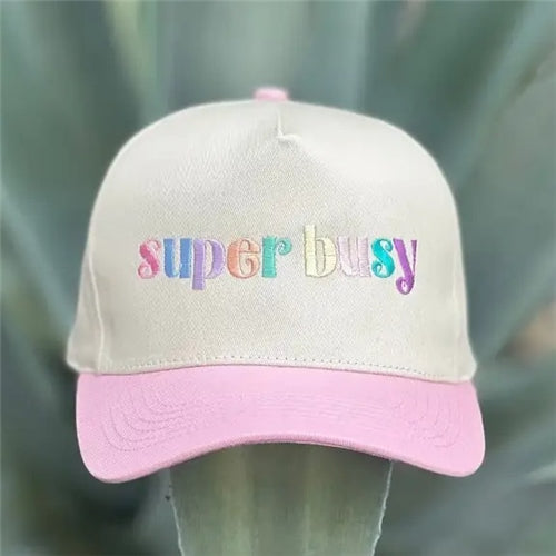 The Super Busy Trucker Hat by Loca on Main features beige embroidery, a pink brim, and "super busy" in colorful pastel letters on the front. This playful hat is shown against a backdrop of blurred greenery.