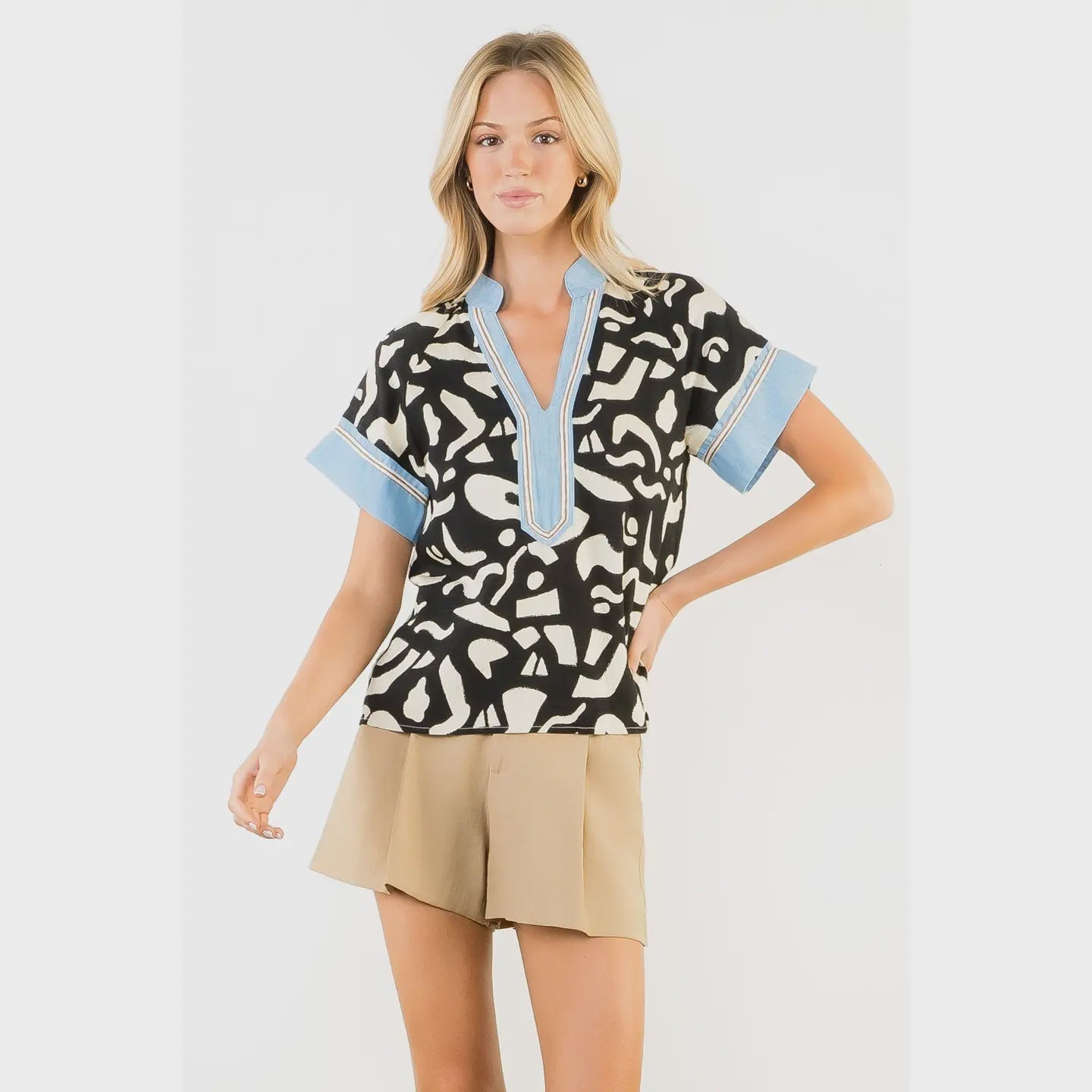 A woman wears the Zertz ABSTRACT PRINT TOP with blue trim and beige shorts, posing confidently against a white background—a chic choice for modern style.