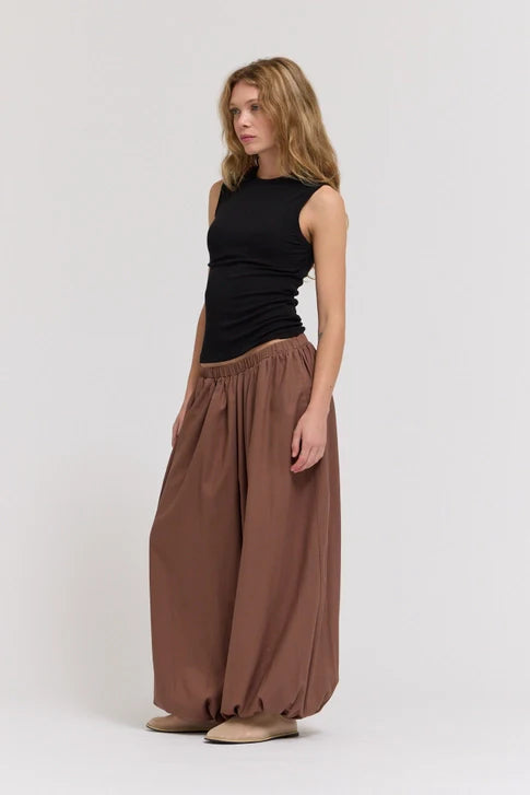 A woman with wavy blonde hair wears the Zertz SHEER TANK TOP, a lightweight sleeveless black top, paired with loose brown balloon pants and beige shoes, standing against a plain light background.
