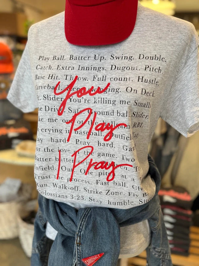 A person wears the Loca on Main "You Play, I Pray Tee," a gray baseball-style shirt with black sayings and bold red script, styled with a red cap and a denim jacket tied around the waist.