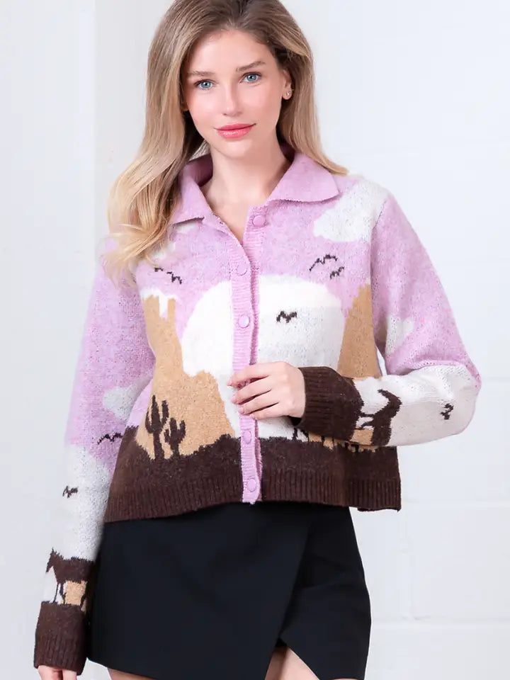 A woman with light brown hair wears the Zertz JESSIE HORSE KNIT SWEATER—a light purple cardigan with a desert landscape, cacti, sun, and birds—paired with a black skirt against a white background.