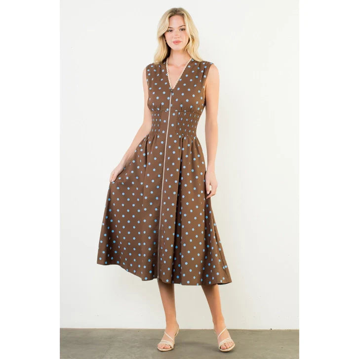 A woman stands against a white wall, wearing the Zertz SLVLESS POLKA DOT MIDI DRESS with light blue spots and a v-neckline. She pairs the lightweight dress with beige sandals and holds its sides while looking slightly off camera.