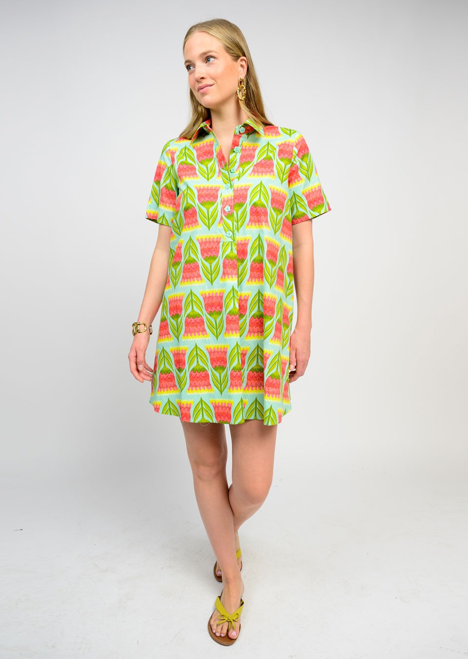 A woman wears the Zertz SWING SHIRTDRESS, featuring bold pink, green, and yellow geometric patterns. She pairs it with gold jewelry and yellow sandals, standing against a plain white background—a versatile wardrobe essential.