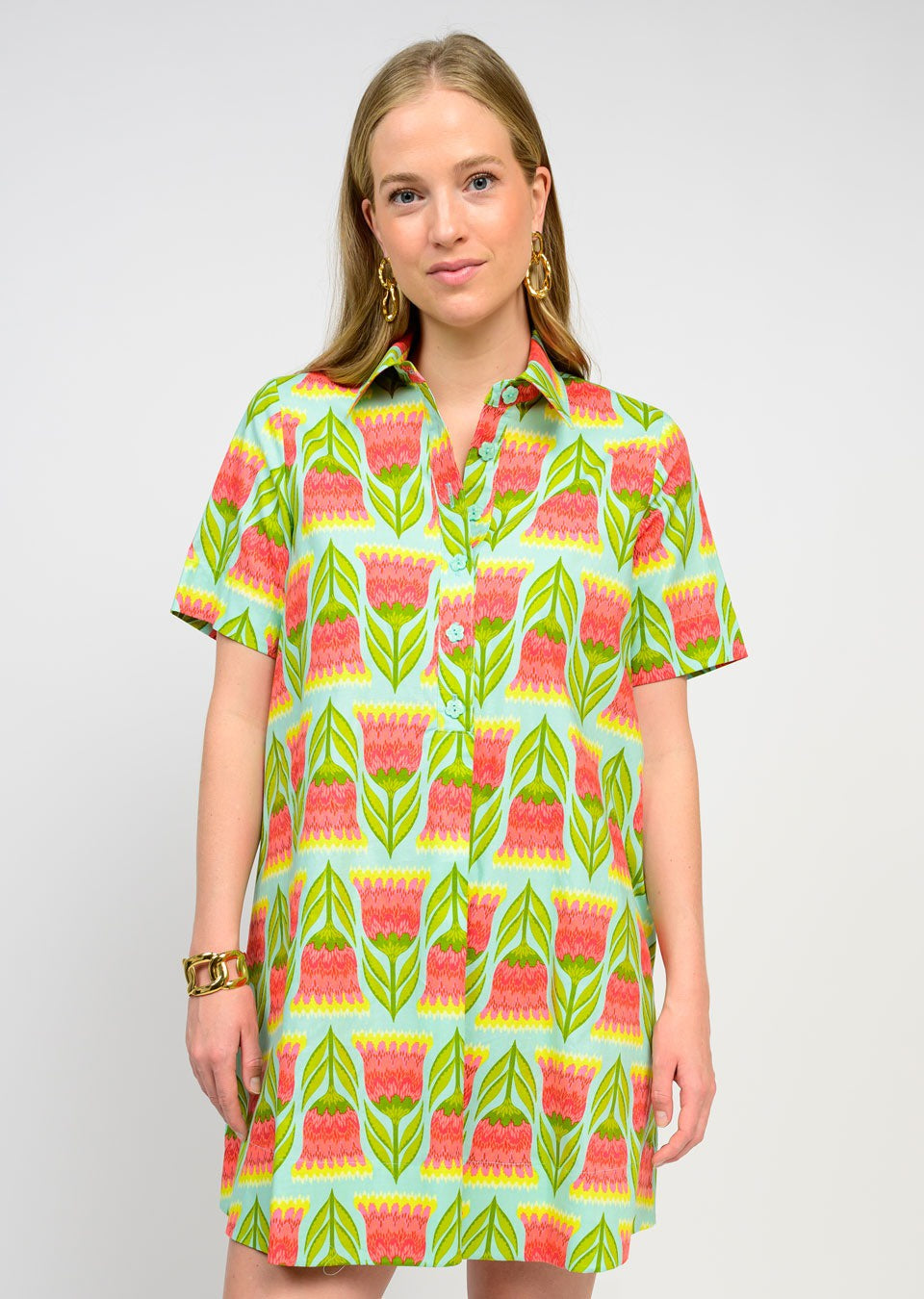 A woman with long blonde hair wears the Zertz SWING SHIRTDRESS, featuring a vibrant green, pink, and orange floral print. Paired with gold earrings and a bracelet, it’s an effortless wardrobe essential against a plain background.