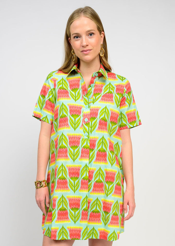 A woman with long blonde hair wears the Zertz SWING SHIRTDRESS, featuring a vibrant green, pink, and orange floral print. Paired with gold earrings and a bracelet, it’s an effortless wardrobe essential against a plain background.