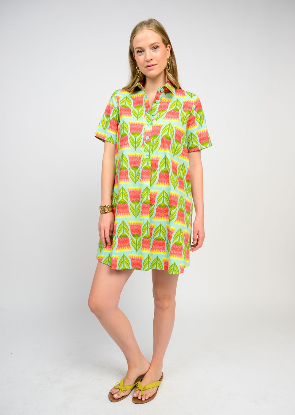 A woman with blonde hair wears the Zertz SWING SHIRTDRESS, featuring short sleeves and a vibrant green, pink, and yellow geometric pattern. She pairs it with gold earrings and yellow sandals for a fresh, essential look.