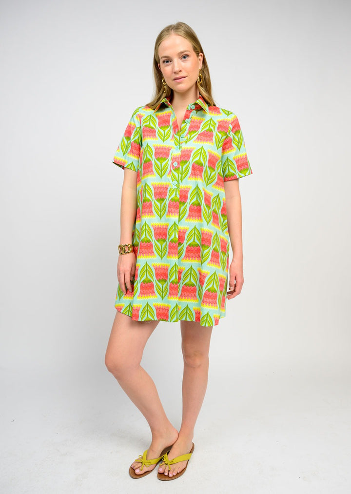 A woman with blonde hair wears the Zertz SWING SHIRTDRESS, featuring short sleeves and a vibrant green, pink, and yellow geometric pattern. She pairs it with gold earrings and yellow sandals for a fresh, essential look.