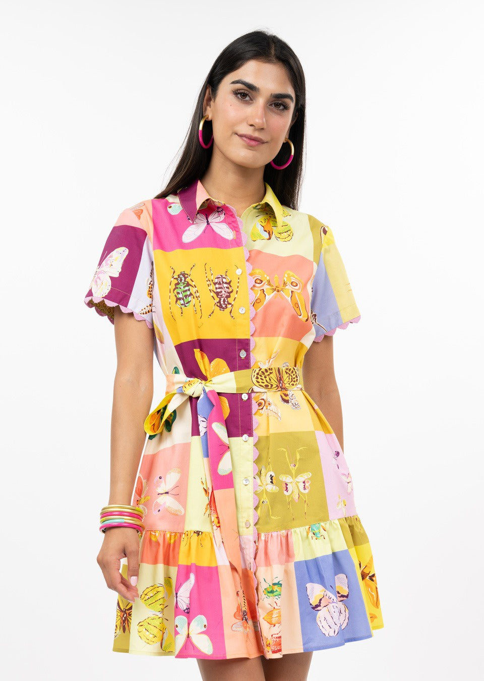 A woman with long dark hair models the Zertz SQUARE DANCE DRESS, a colorful, short-sleeved, button-down dress with a butterfly and patchwork pattern. She pairs it with pink hoop earrings and bracelets against a white background.