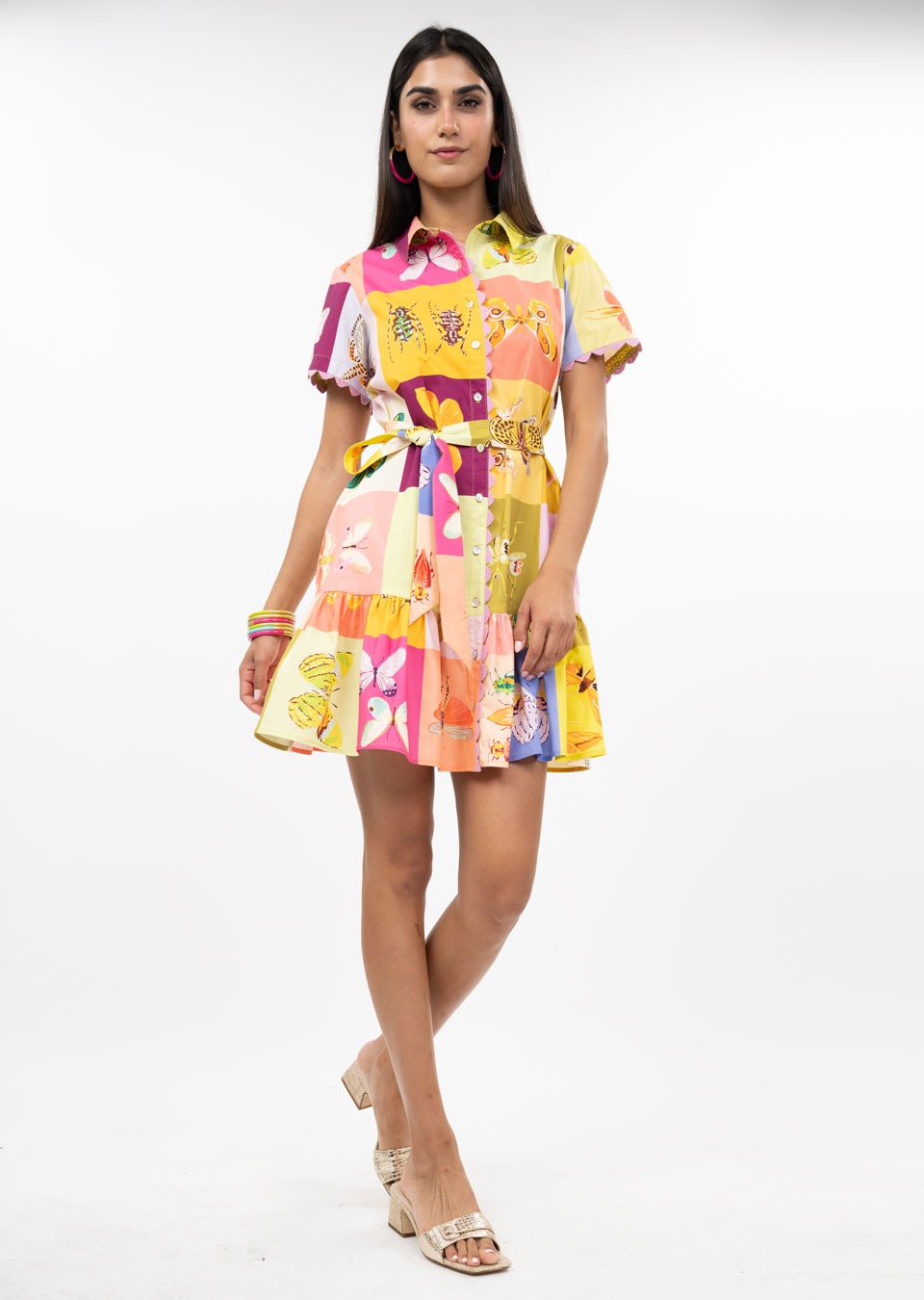 A woman models the Zertz SQUARE DANCE DRESS, featuring a vibrant floral and geometric pattern, short sleeves, and a button front. She pairs the dress with sandals and accessories, twirling against a white background.