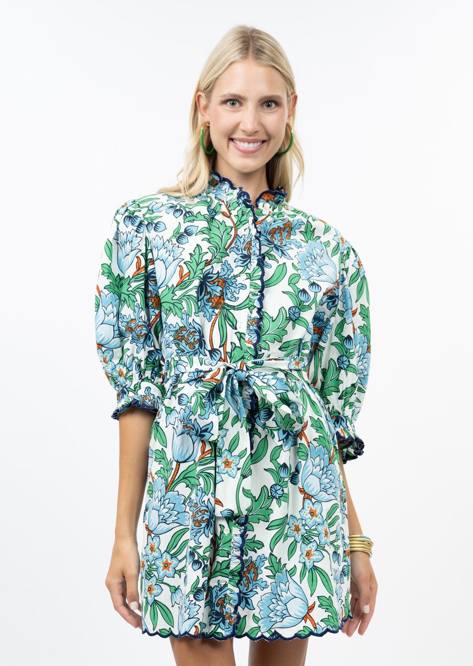 A woman with blonde hair and hoop earrings smiles at the camera against a white background, wearing the Zertz SCALLOP FRONT DRESS featuring blue scalloped edges, puffed sleeves, and a floral belt with green, blue, and orange flowers.