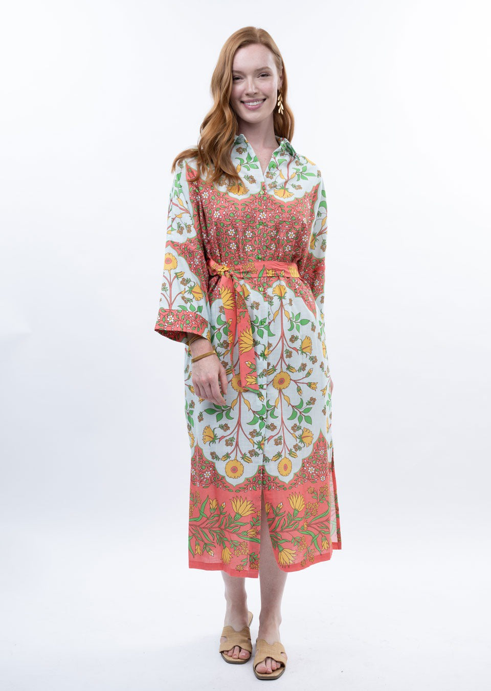 A woman with long red hair smiles in the Zertz BORDERED SHIRT DRESS, a colorful floral piece with a belted waist and long sleeves. She stands against a plain white background, paired with open-toed sandals.