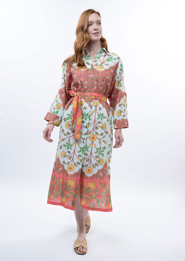 A woman with long red hair wears the Zertz BORDERED SHIRT DRESS, featuring long sleeves, a belted waist, and pink, yellow, and green floral designs. She looks to the side in beige sandals against a white background.