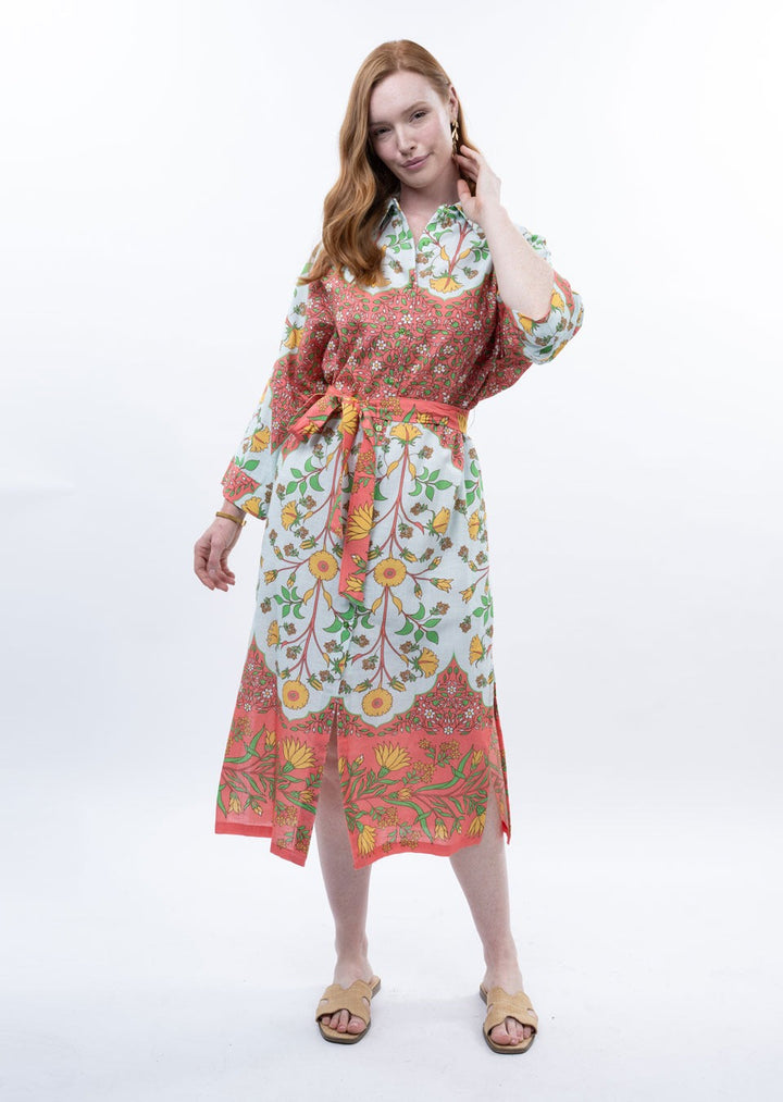 A woman with long, wavy red hair wears the Zertz BORDERED SHIRT DRESS—a colorful shirt dress with a waist tie and floral border. She stands on a white background, smiling slightly, and pairs it with flat beige sandals.