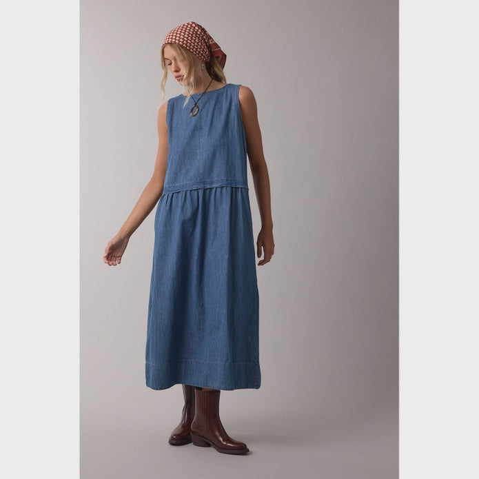 A woman models the SLVLESS MAXI DRESS by Zertz—an ankle-length, sleeveless denim style—paired with brown boots and a red patterned headscarf, standing against a light gray background with one arm slightly outstretched.