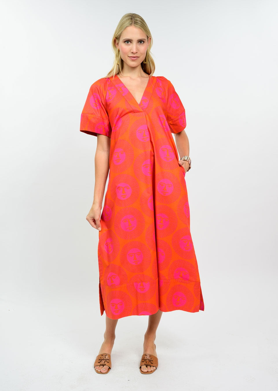 A woman with blonde hair poses barefoot against a white background, wearing the Zertz HERE COMES THE SUN DRESS—a vibrant midi with orange and pink patterns, short sleeves, and a V-neck—smiling gently.