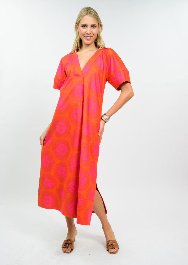 A woman smiles in the Zertz HERE COMES THE SUN DRESS—a red and pink patterned V-neck midi with short sleeves and pockets—paired with brown sandals and a watch, standing against a plain white background.