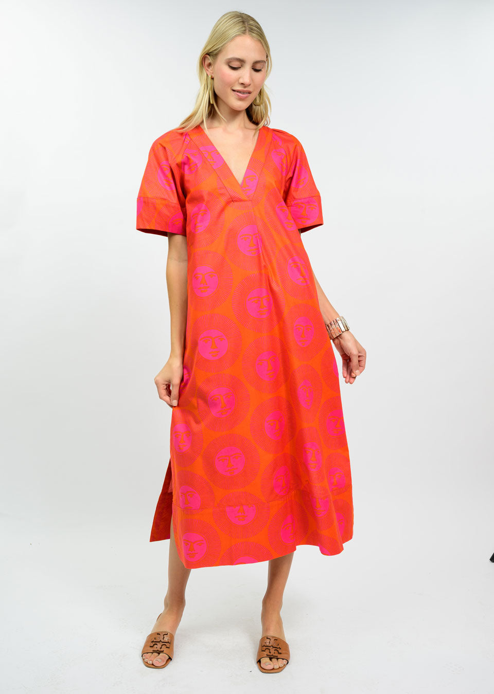 A woman with blonde hair poses against a white background wearing the Zertz HERE COMES THE SUN DRESS—a loose-fit, red and pink abstract midi—styled with brown sandals and a watch.