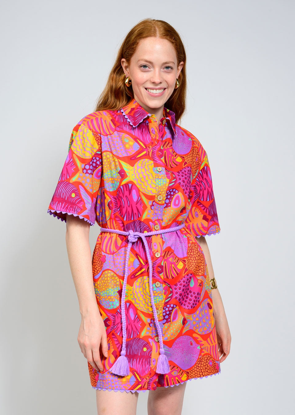 A smiling woman with long red hair models the Zertz SCHOOL OF FISH DRESS, featuring a vibrant pink, orange, and purple fish print, short sleeves, belted waist, and scalloped trim. She stands against a plain light background.